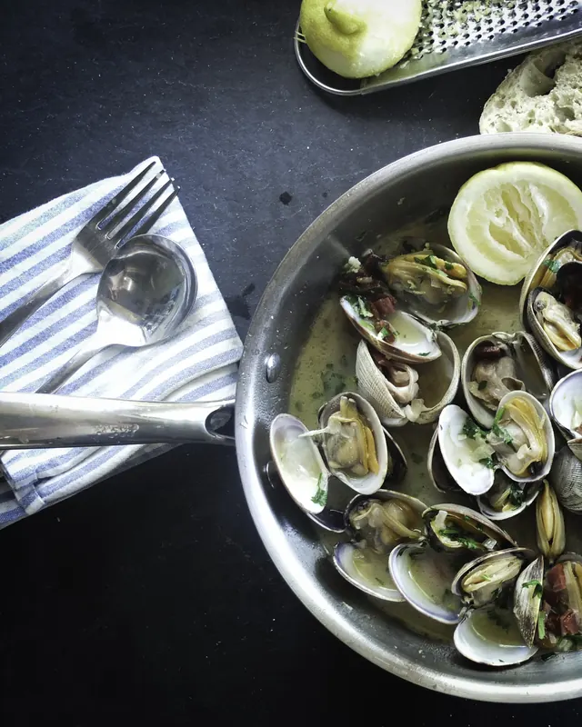 Clams opened in a steel pan with lemon, parsley and a spoon set beside it.